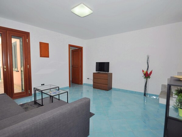 A living room with a grey sofa, a glass coffee table, a wooden TV stand, and a TV. There is a door leading to another room, and a potted plant in the corner. - Appartamento a Cetara ID 722 (Cetara)