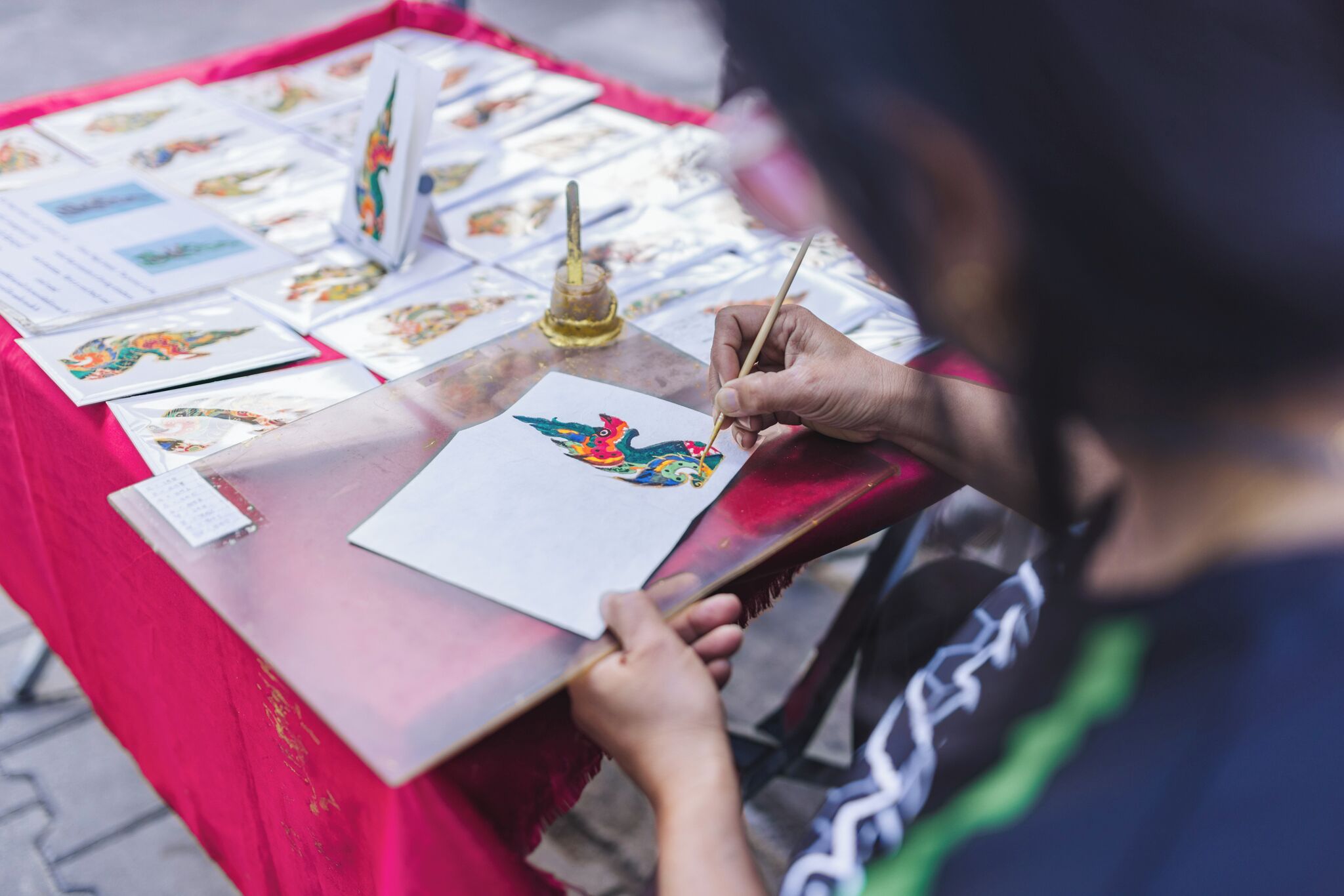Una persona pintando sobre una mesa con varias obras de arte y una pequeña botella.