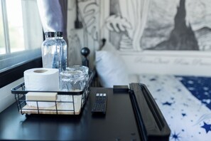 A room with a bed, a window, a bedside table with a remote control, a tissue roll, and two water bottles.