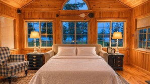 A wooden cabin bedroom with a large bed, two nightstands, a chair, and a view of trees through the windows. - Charlevoix Lakefront Cottage (Charlevoix)