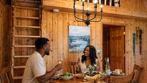 A wooden interior with a dining table, chairs, a chandelier, and a painting of a landscape. - Charlevoix Lakefront Cottage (Charlevoix)