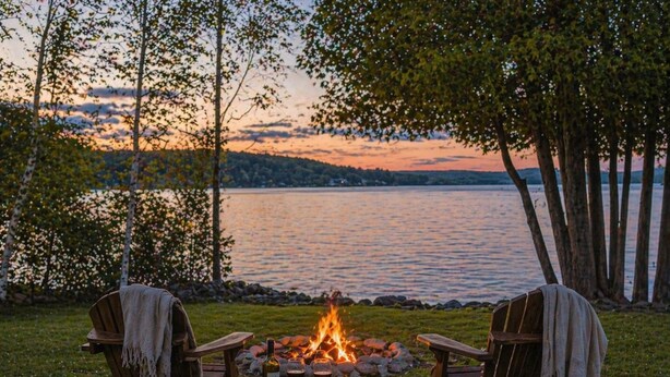 Two wooden chairs with blankets, a small round table with a bottle and glasses, and a fire pit with rocks in a grassy area with trees and a lake in the background. - Charlevoix Lakefront Cottage (Charlevoix)