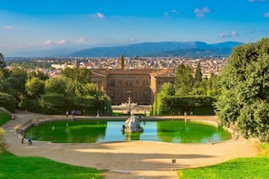 A historic building with a fountain in a park, surrounded by greenery and a cityscape in the background.
