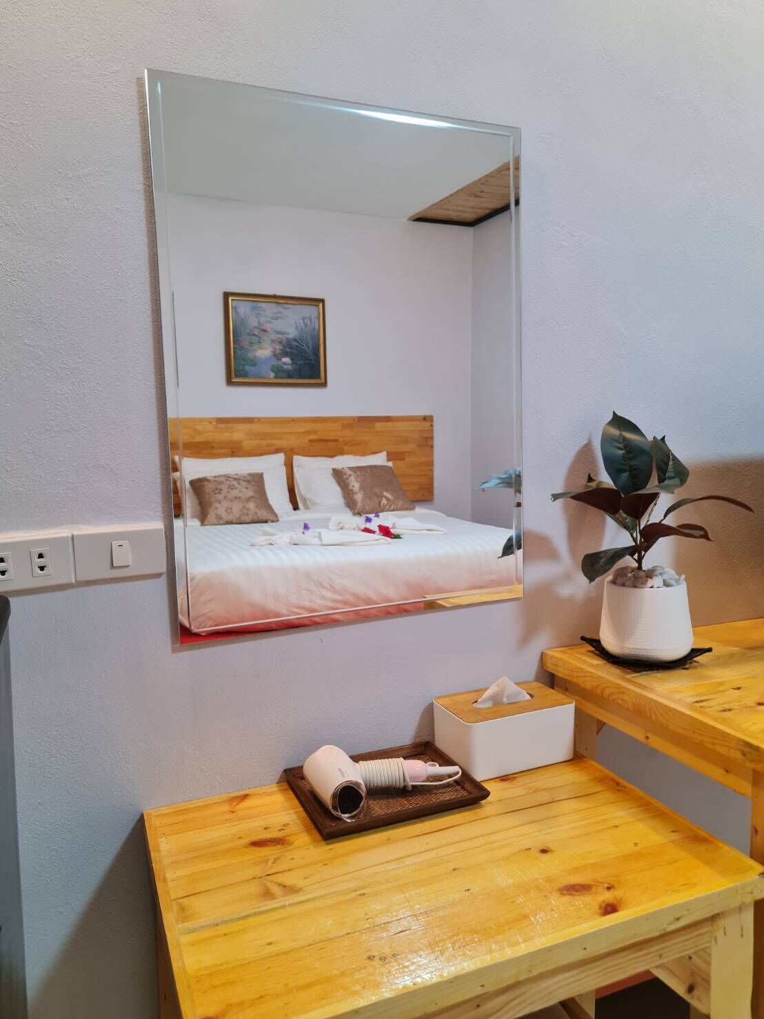 A bedroom with a bed, a bedside table with a hairdryer, a mirror, and a potted plant.