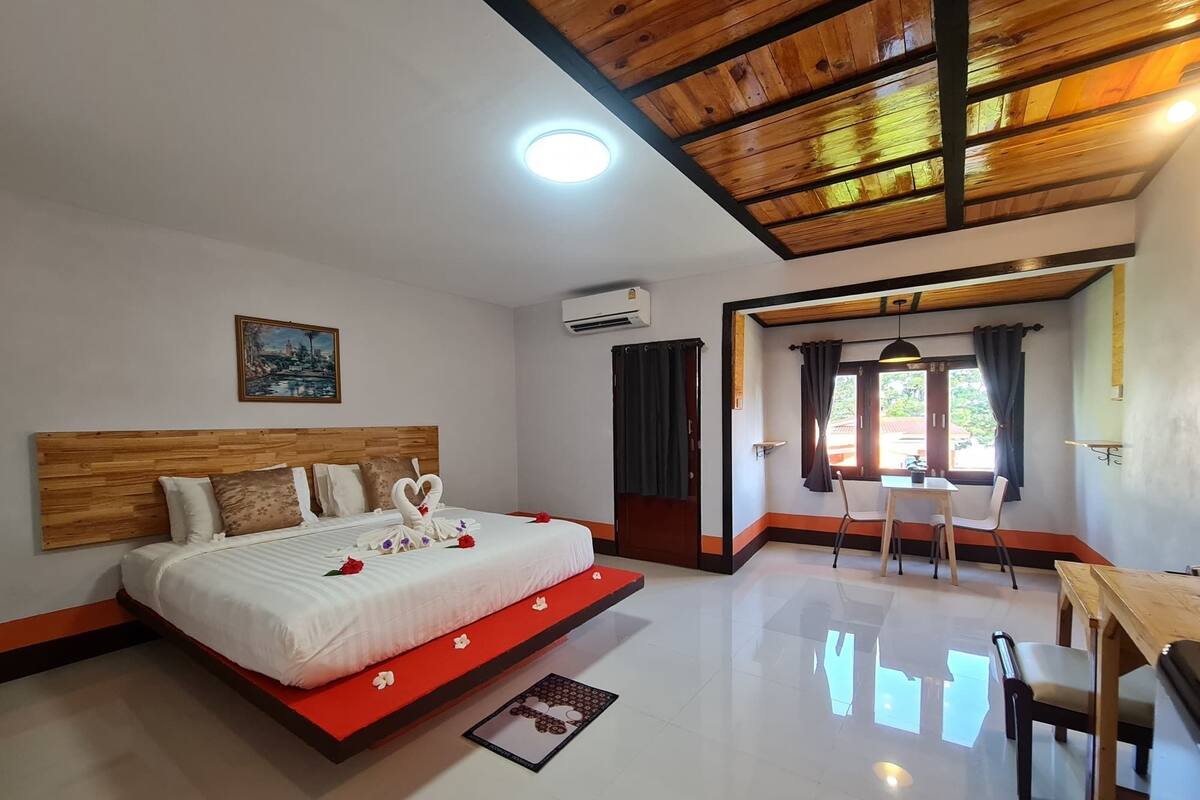 A hotel room with a wooden ceiling, a bed with a red runner, a dining table with chairs, and a painting on the wall.