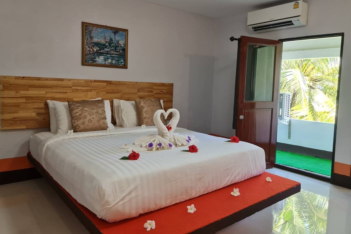 A hotel room with a wooden headboard, a bed with white linens, a painting on the wall, and a view of palm trees through a glass door.