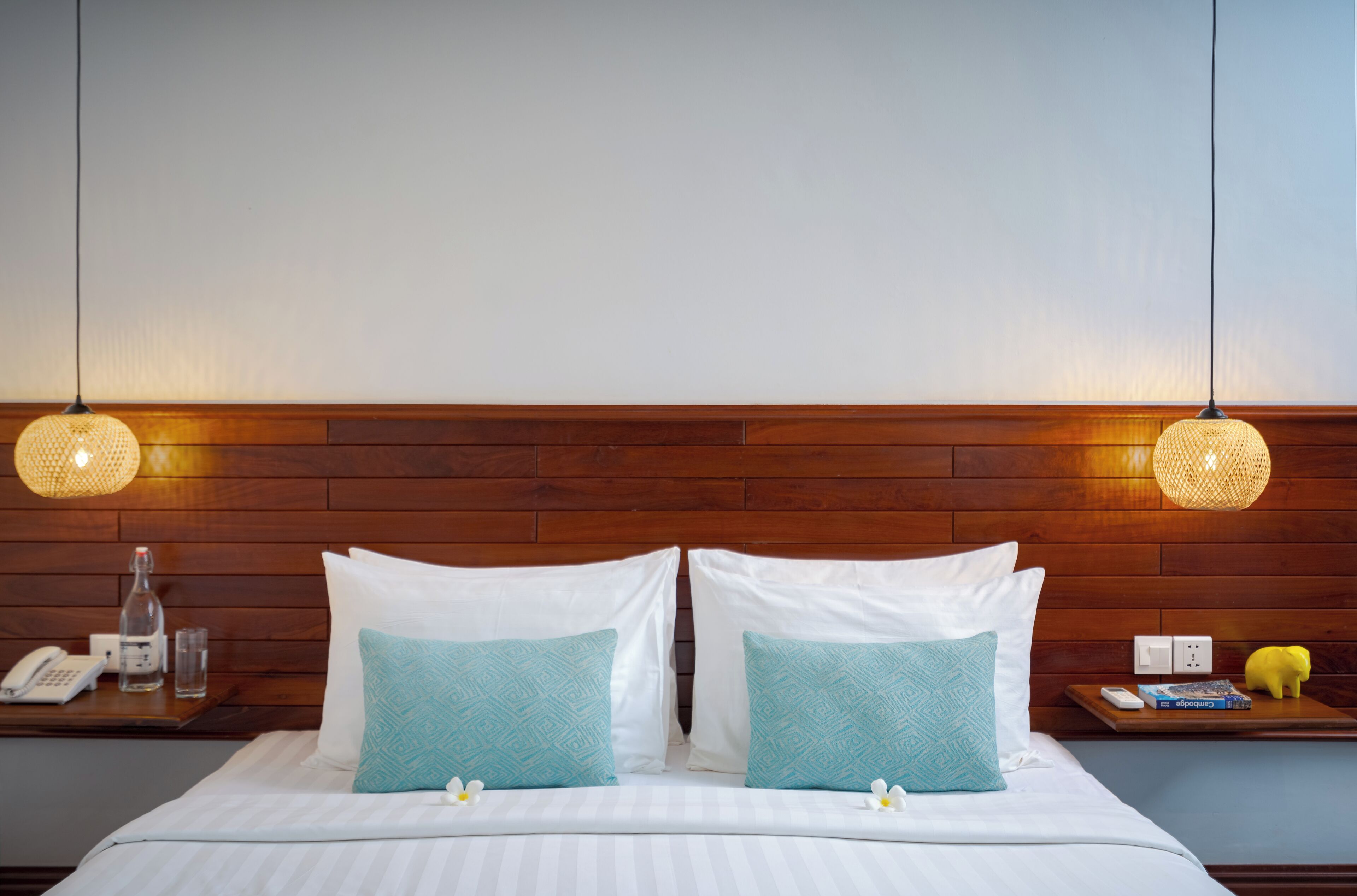 Un letto rifatto con cuscini bianchi e blu, un comodino con un telefono e un bicchiere, e una testiera in legno.