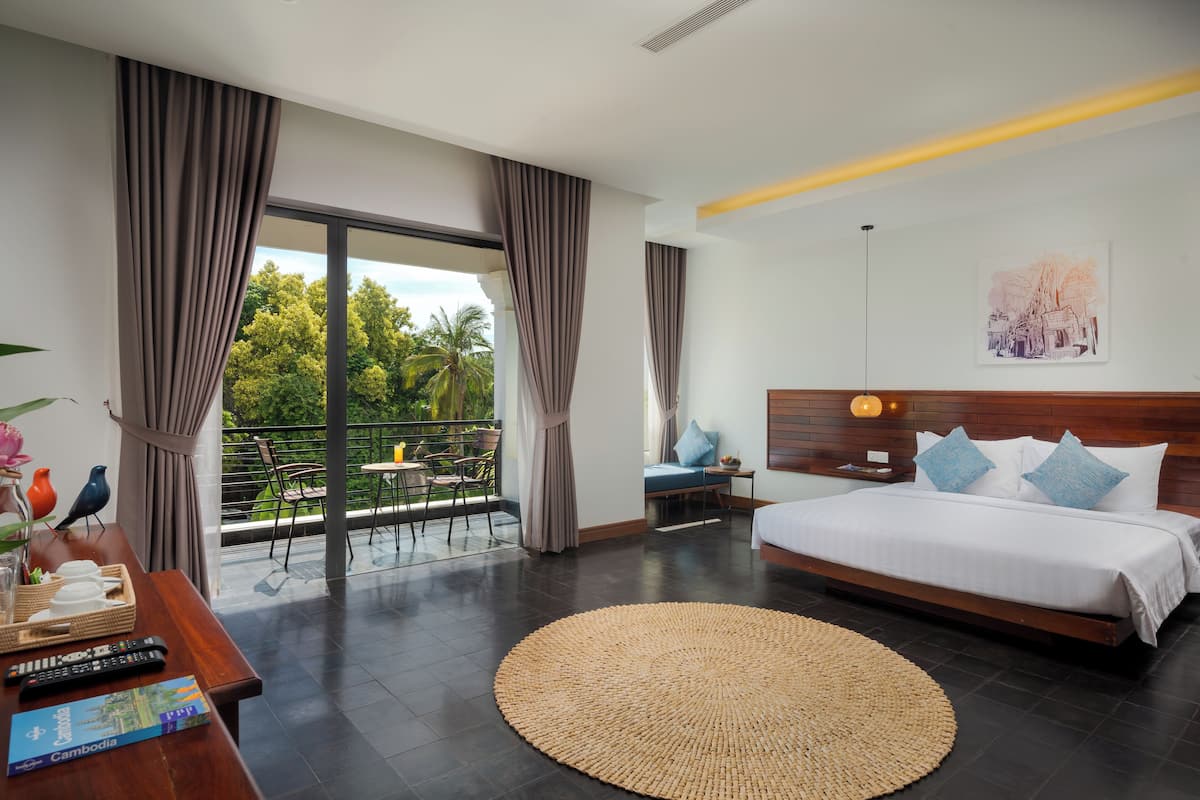Ein modernes Hotelzimmer mit einem großen Bett, einem Balkon mit Blick auf Grünflächen, einem Essbereich mit Tisch und Stühlen sowie einem hölzernen Kopfteil mit einer Lampe.