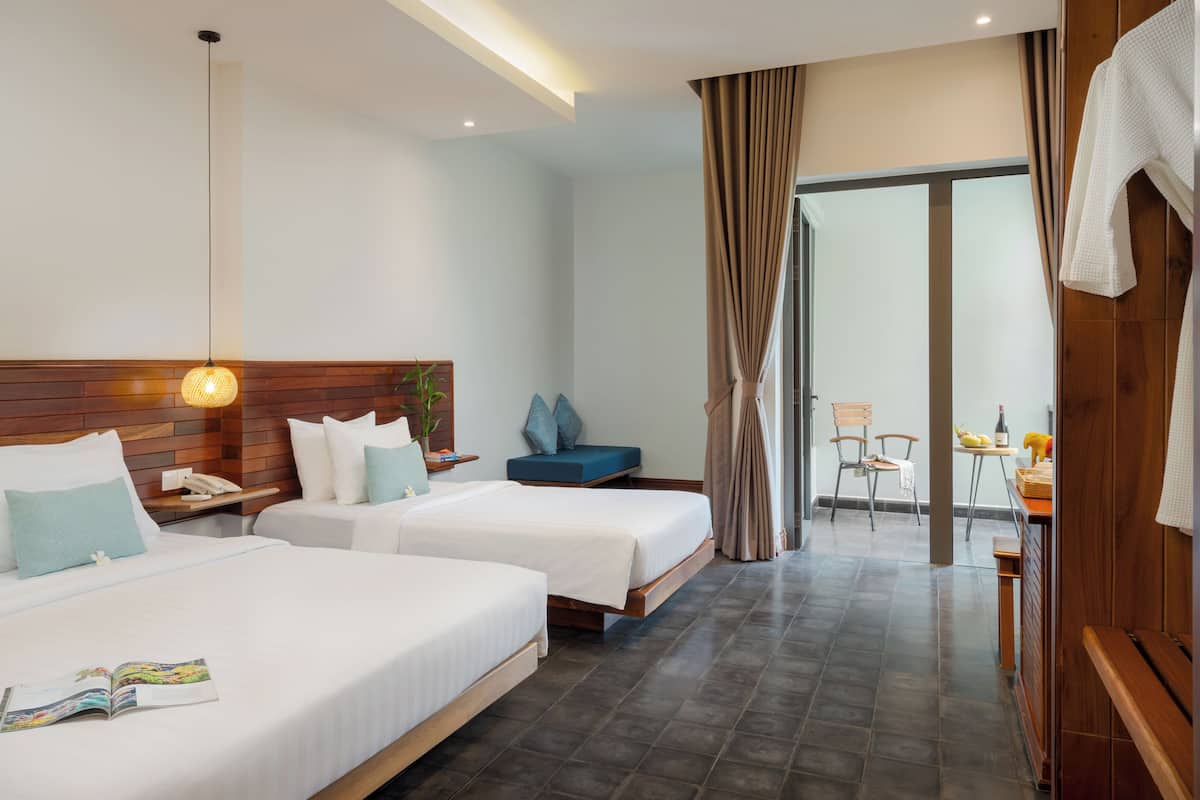 Ein Hotelzimmer mit zwei Betten, einem Holzkopfende, einem Nachttisch mit Lampe und Blick auf den Speisesaal durch eine Glastür.