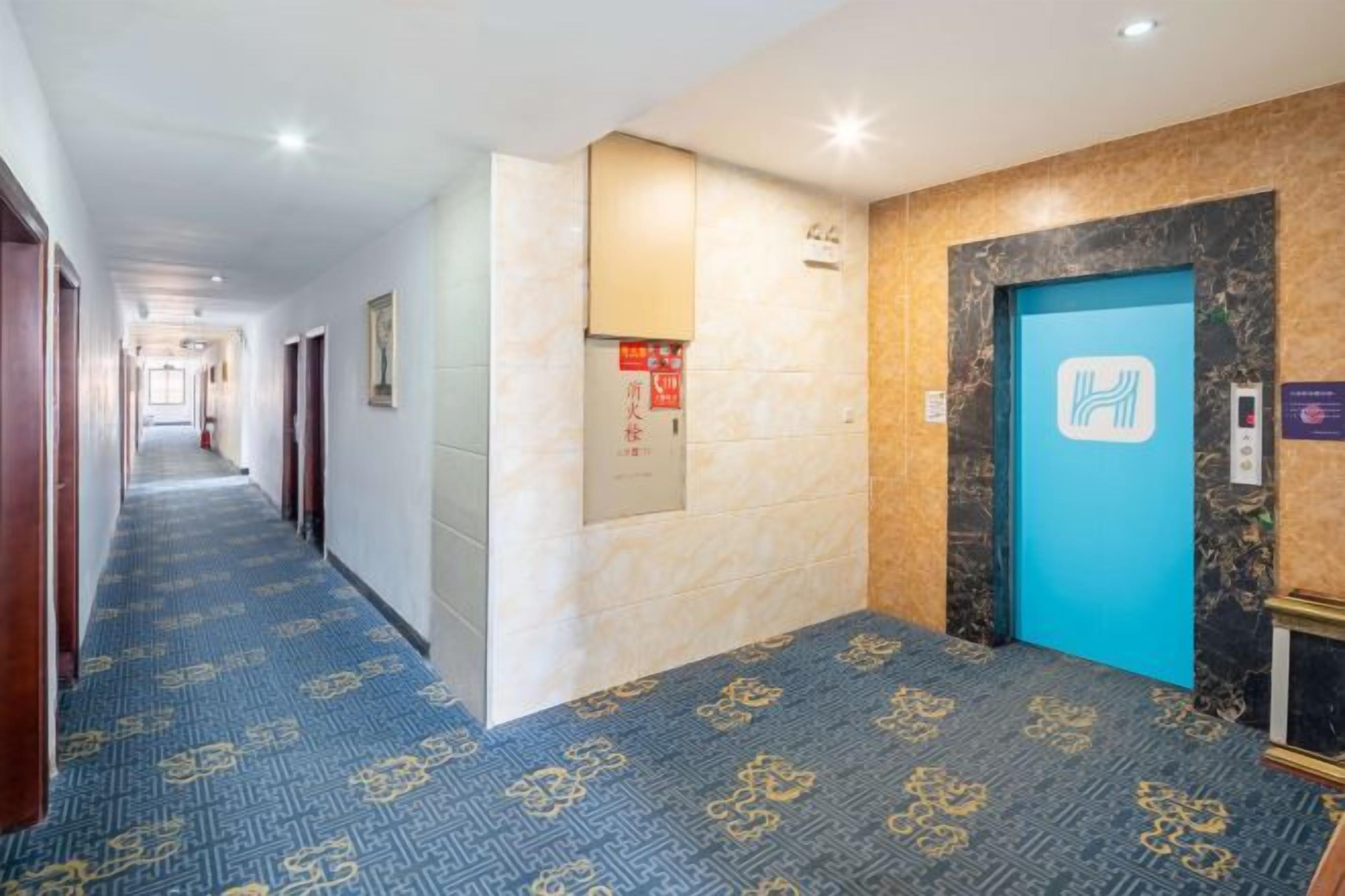 Pasillo de hotel con alfombra estampada, puerta de ascensor azul y una zona de recepción con un cartel.