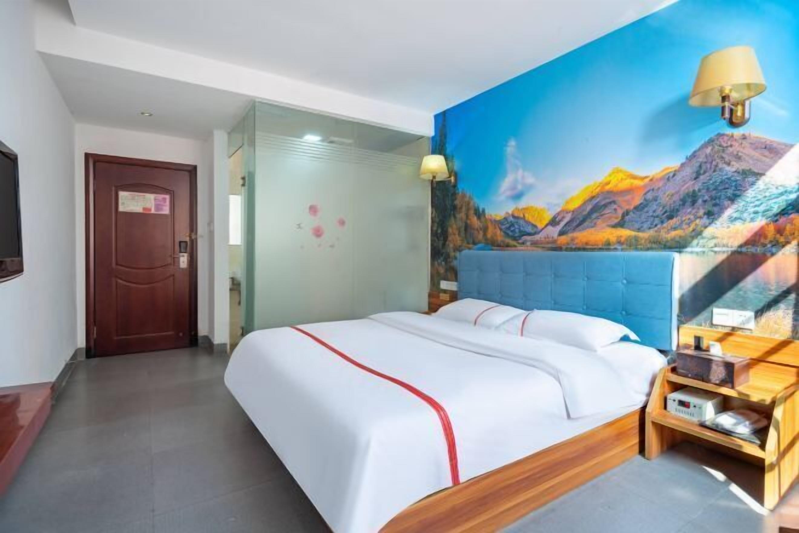 A hotel room with a bed, a wooden nightstand, a wall-mounted lamp, and a painting of a mountain landscape.