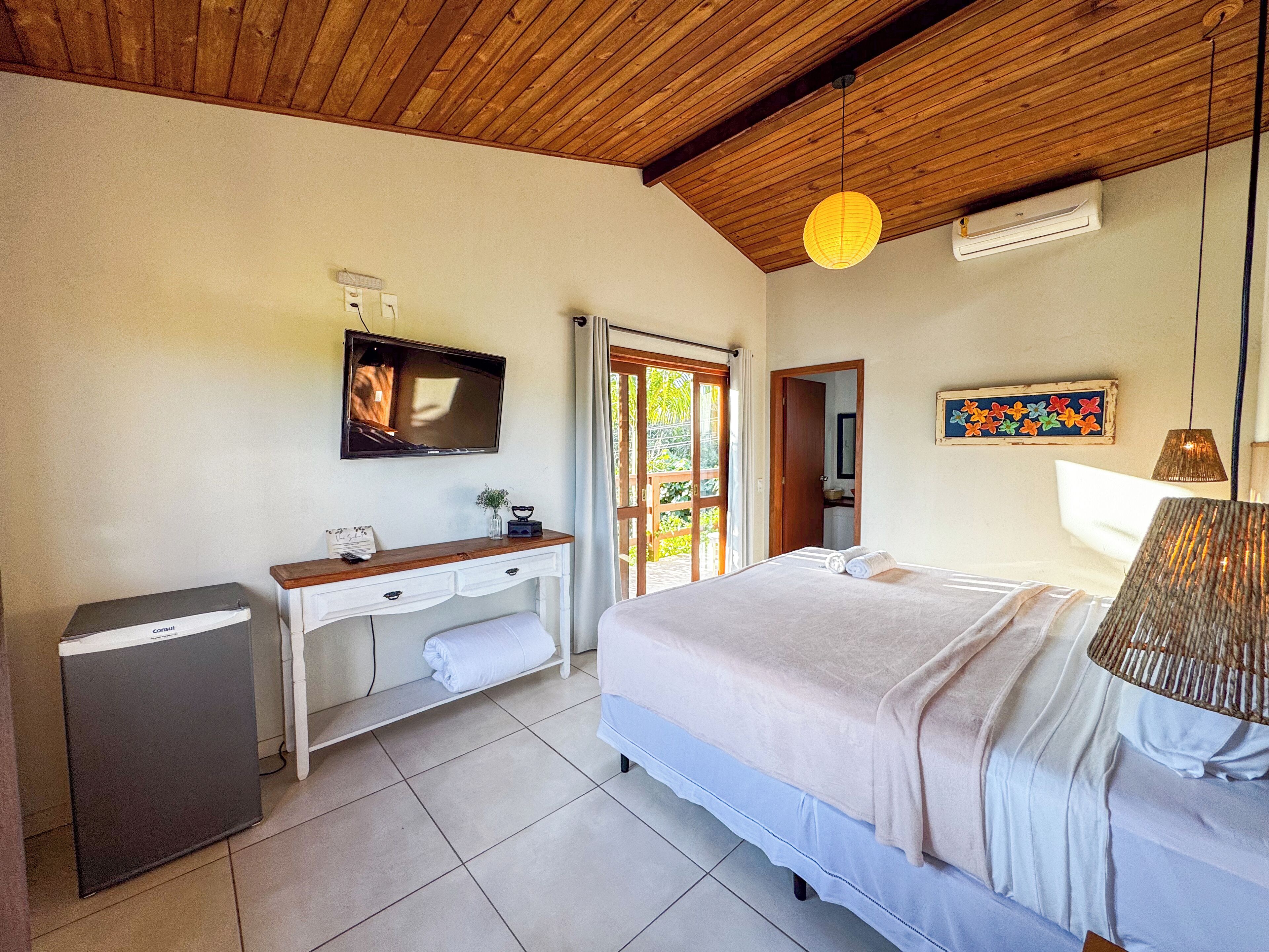 A bedroom with a bed, a desk with a television, a small trash bin, and a door leading to a balcony.