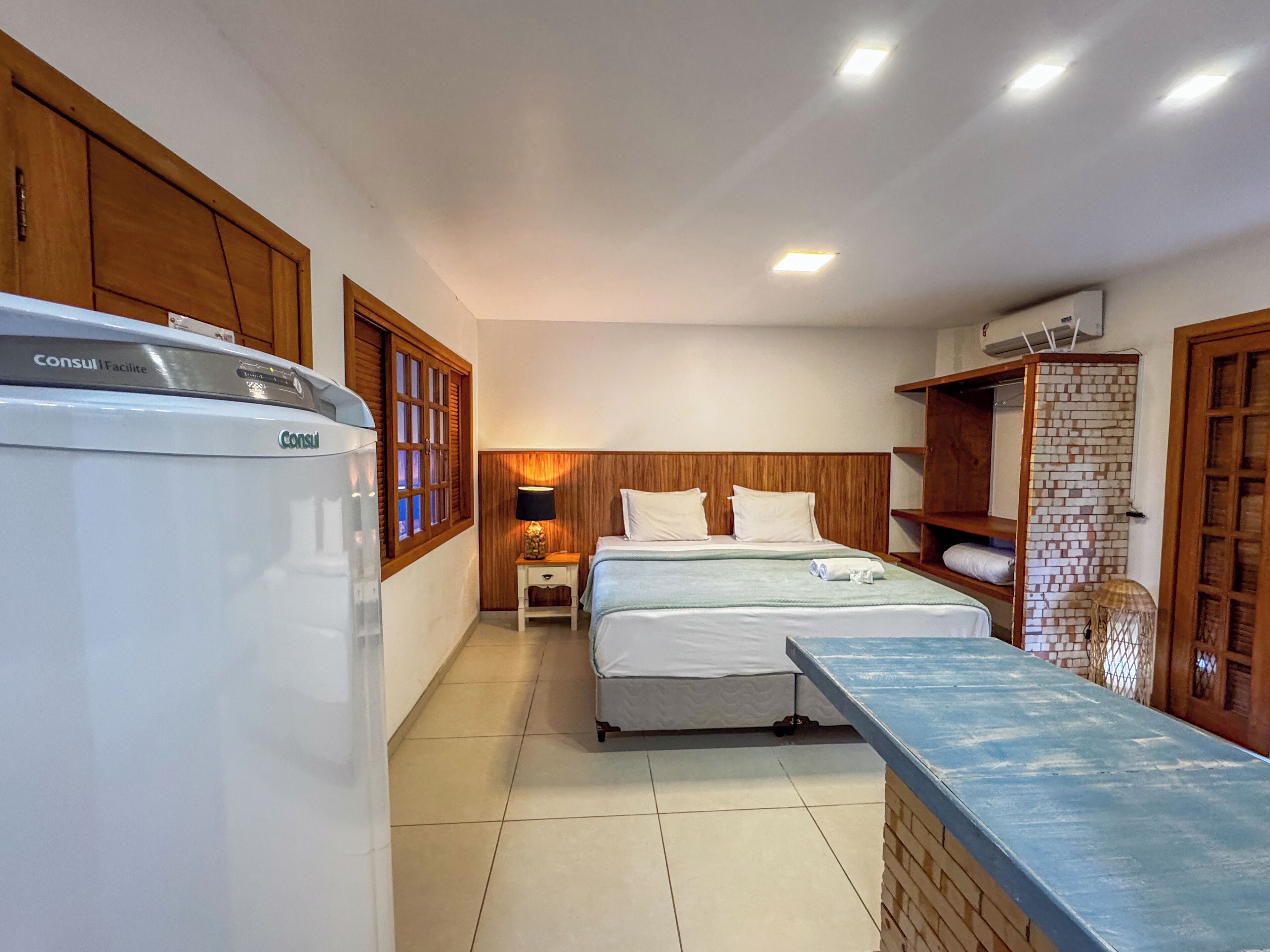 Habitación de hotel con cama, mesita de noche con lámpara, armario de madera y kitchenette con refrigerador.