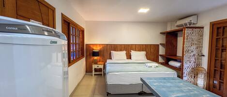 Habitación de hotel con cama, mesita de noche con lámpara, armario de madera y kitchenette con refrigerador.
