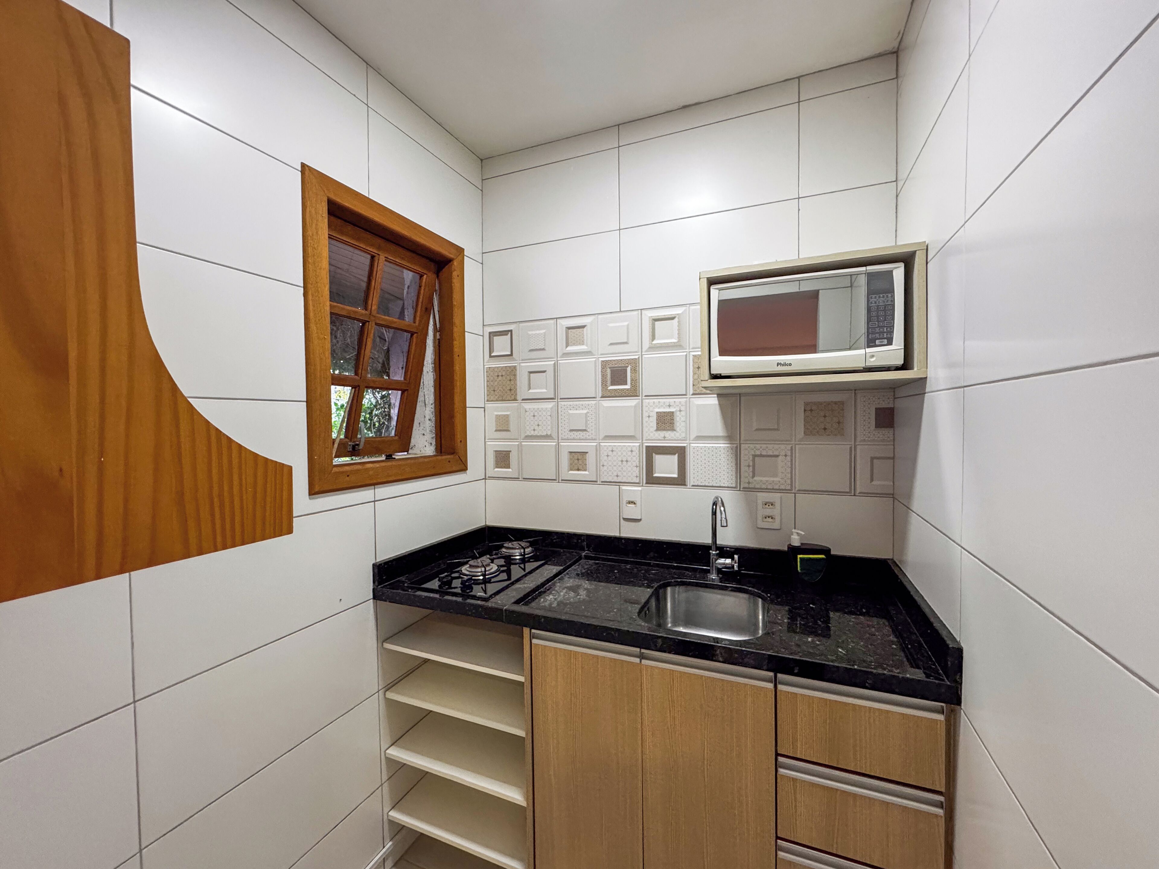 Una cocina compacta con baldosas blancas, encimera negra, estufa integrada, un lavamanos y armarios de madera.