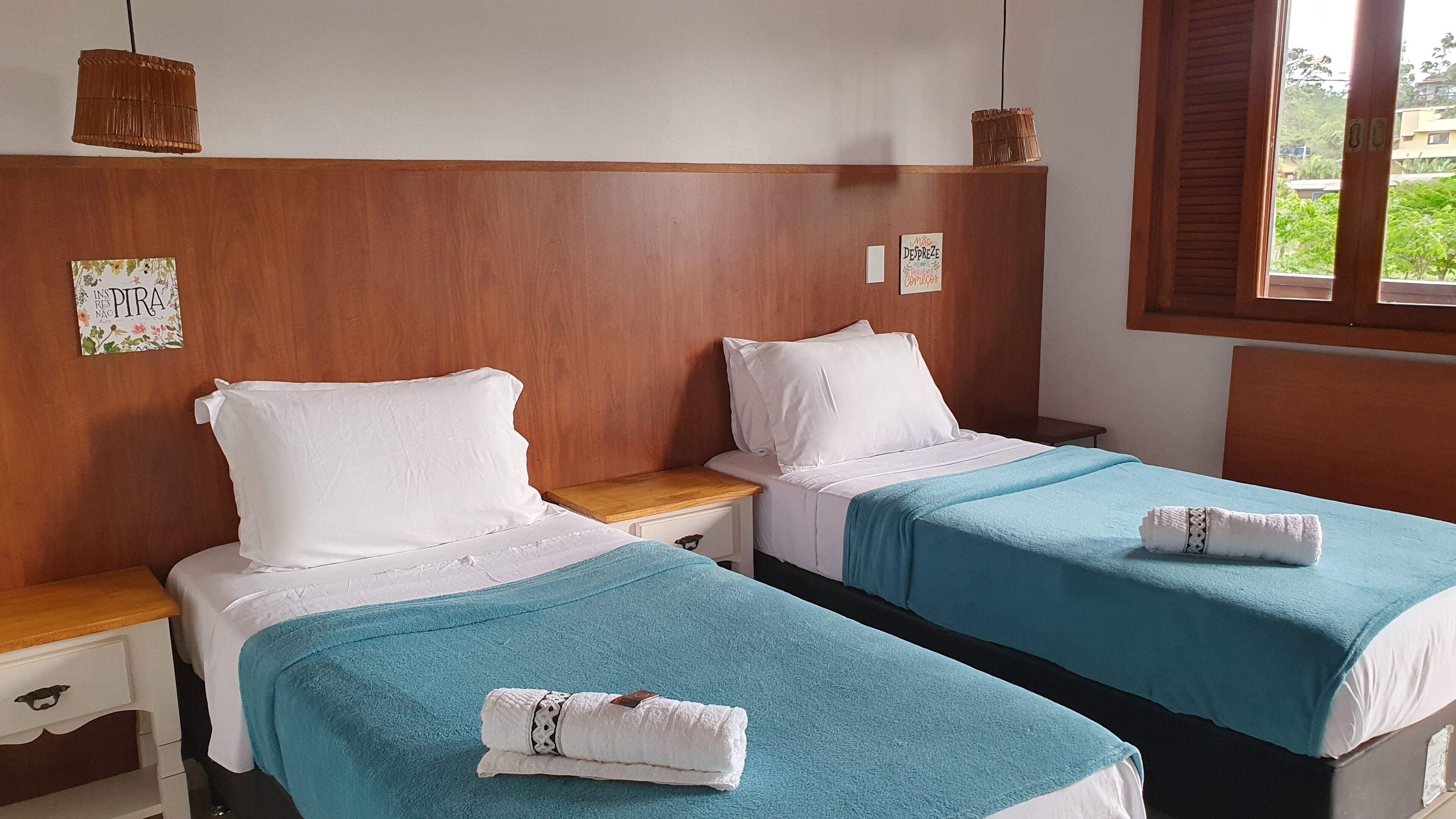 Una habitación con dos camas individuales, cabeceros de madera, una mesita de noche de madera, un cajón blanco y una ventana con vistas a la vegetación.