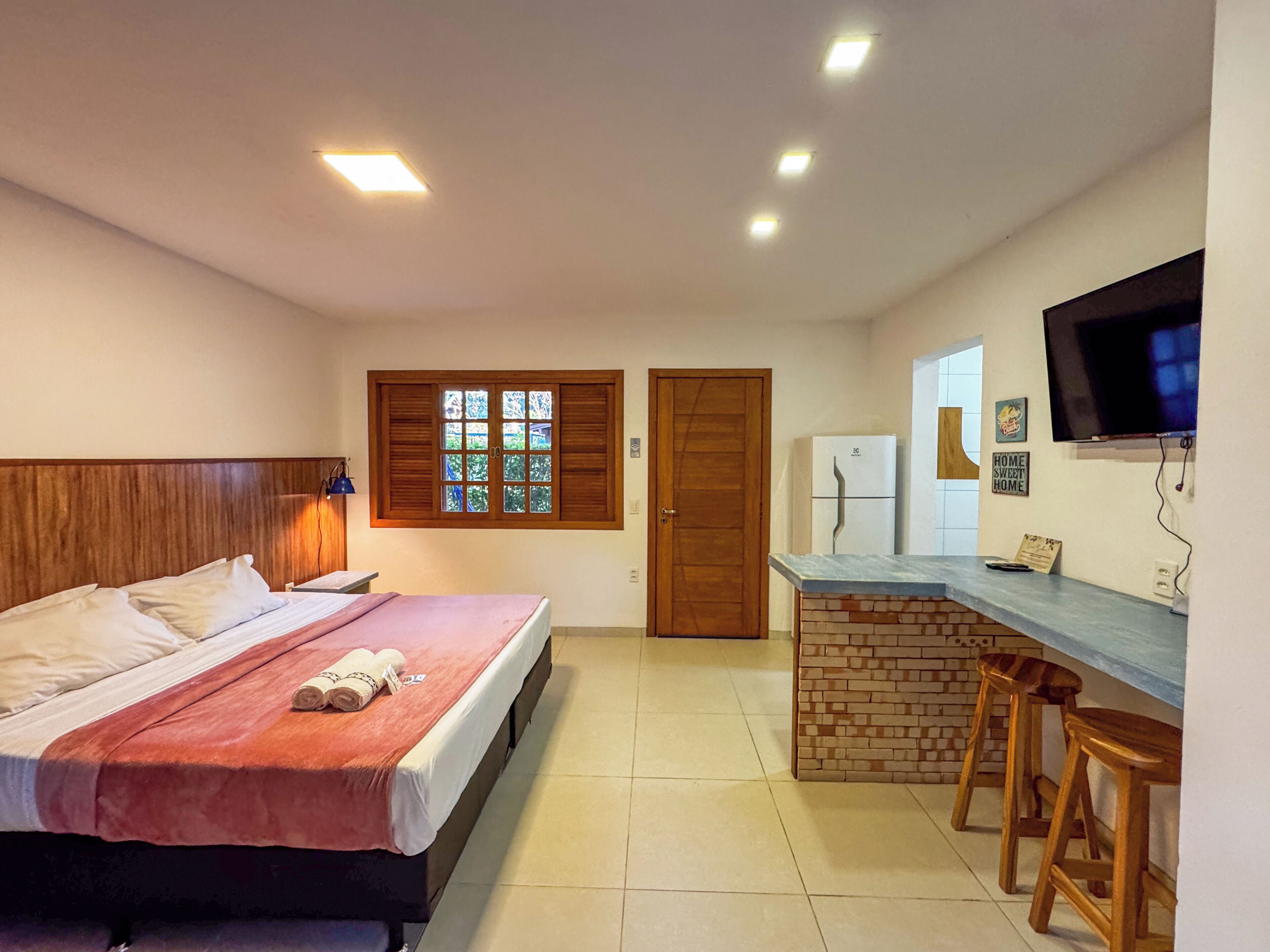 A hotel room with a bed, a small kitchenette, a TV, and a window with shutters.