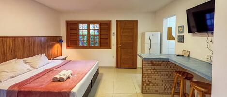 A hotel room with a bed, a small kitchenette, a TV, and a window with shutters.