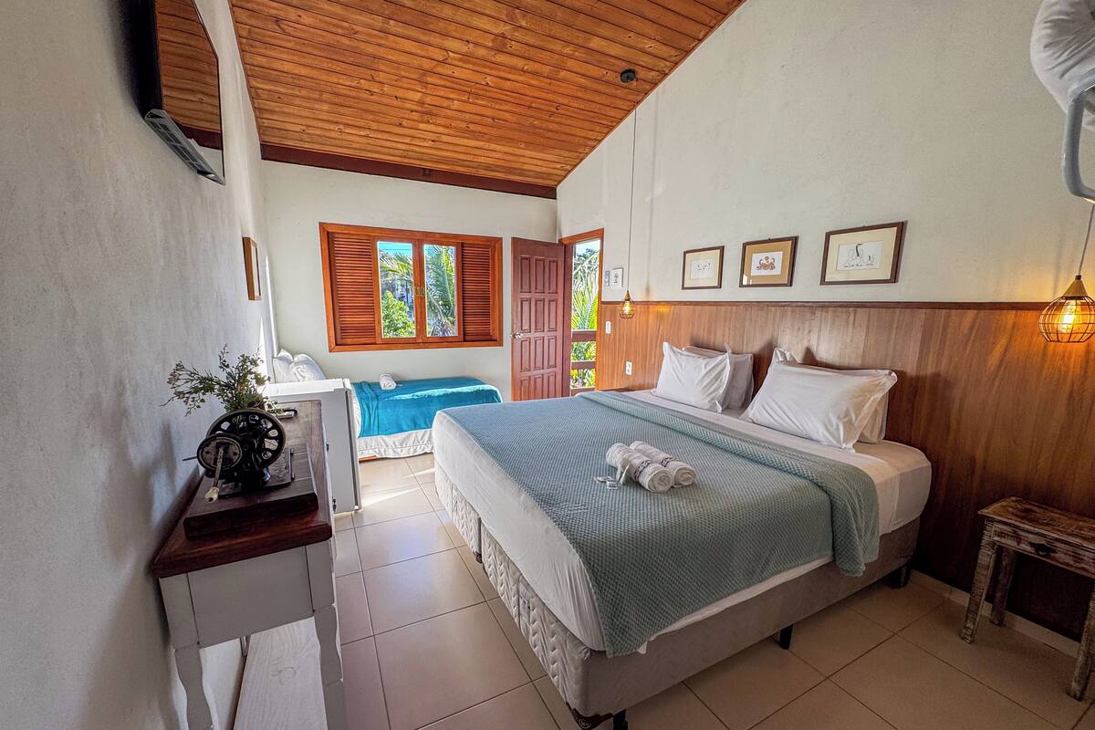 A bedroom with a large bed, a desk with a plant, and a view of the outdoors through a window with shutters.