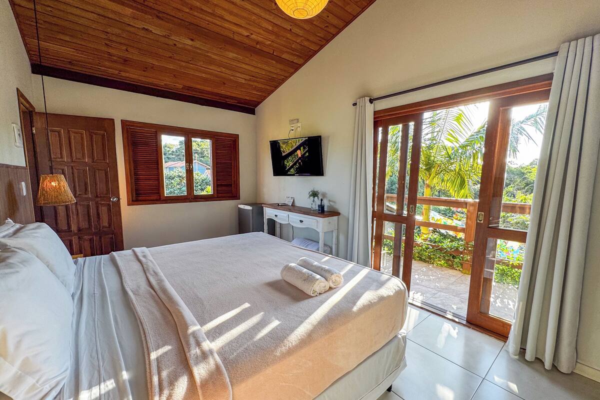 A bedroom with a large bed, a desk, a TV, and a sliding glass door to a garden.