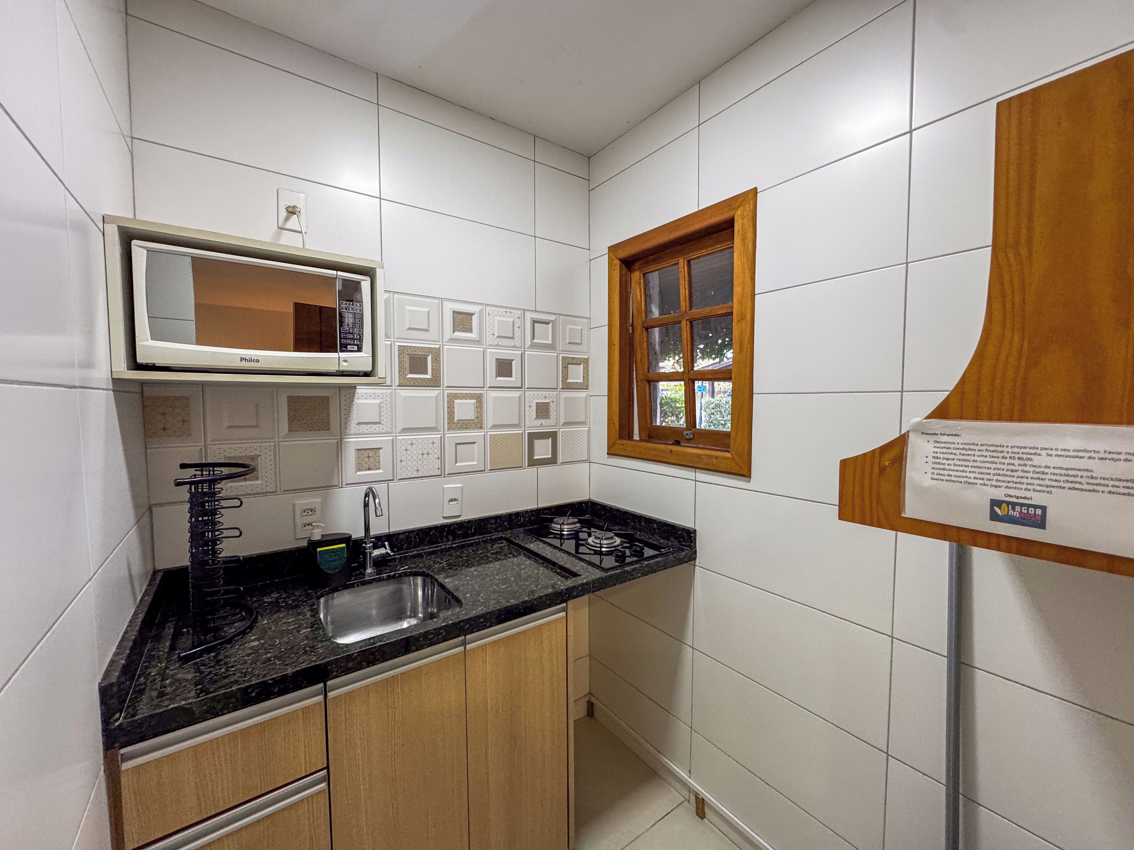 A compact kitchen with white tiles, a black countertop, a built-in microwave, a sink, and wooden cabinets.