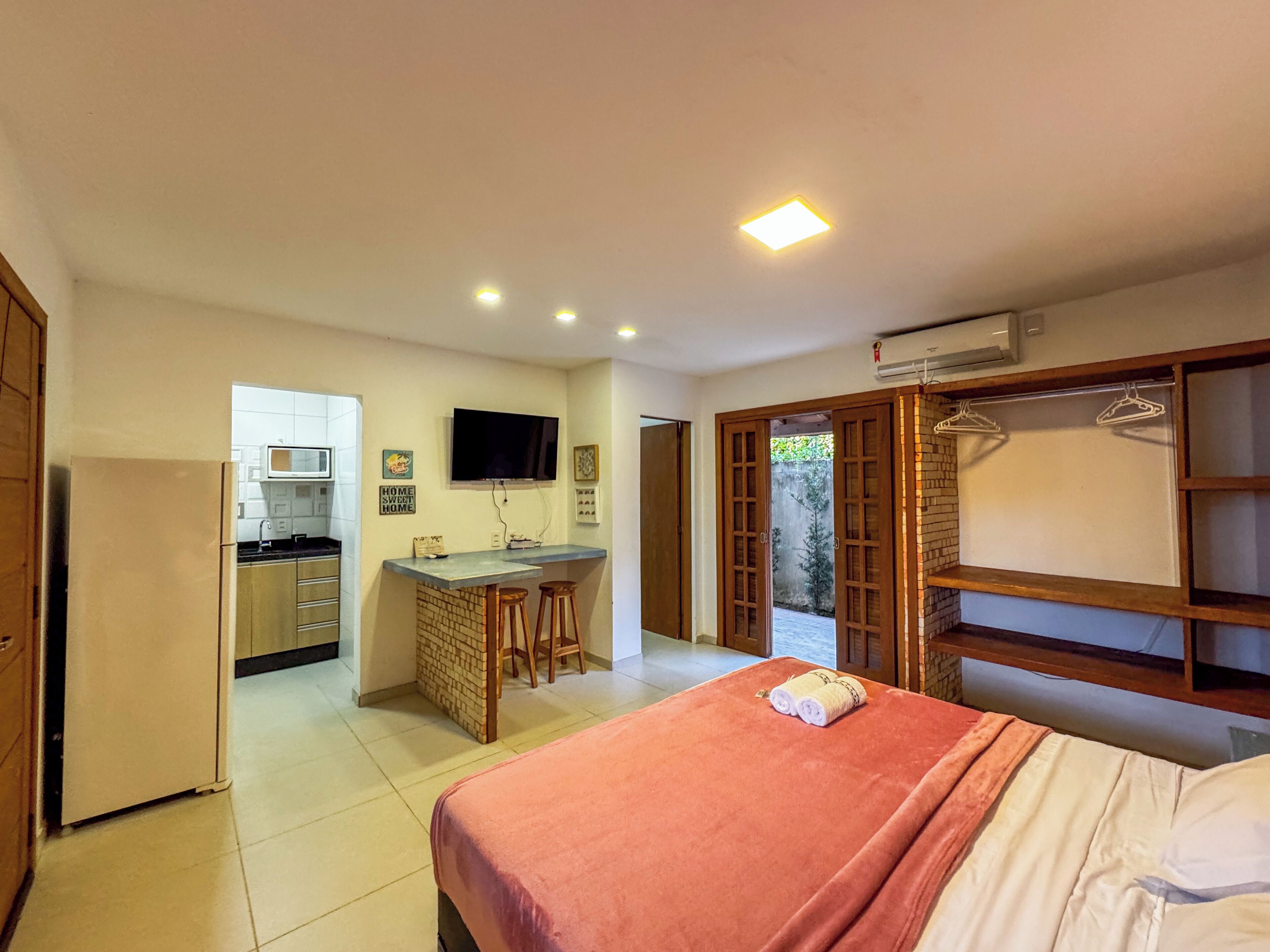 Una habitación de hotel compacta con kitchenette, una cama con una colcha roja y un armario de madera con perchero.