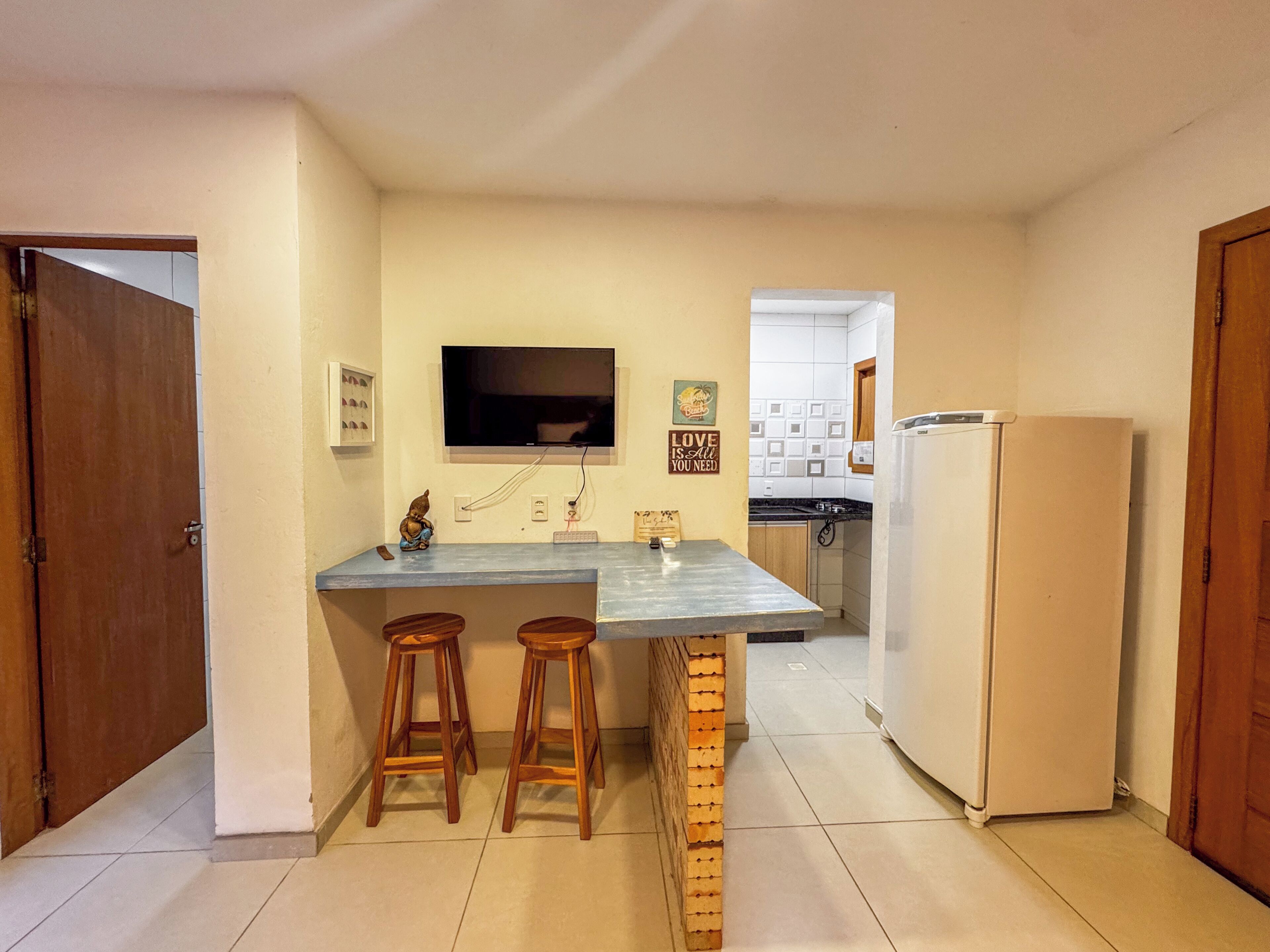 A compact kitchen with a central island, wooden stools, a refrigerator, and a television mounted on the wall.