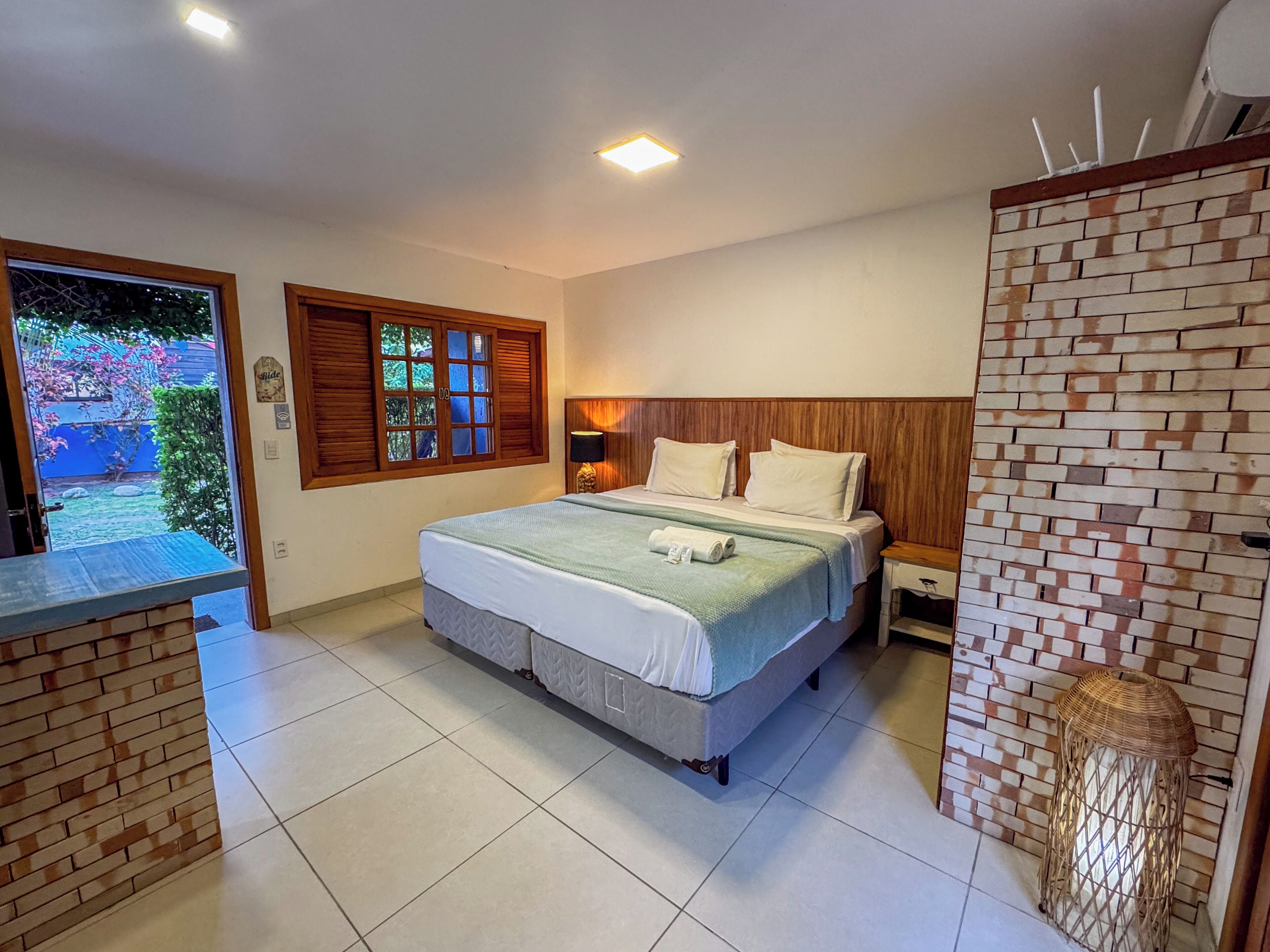 Ein Hotelzimmer mit einem großen Bett, einem kleinen Nachttisch, einem Fenster mit Rollläden und Blick auf einen Garten.