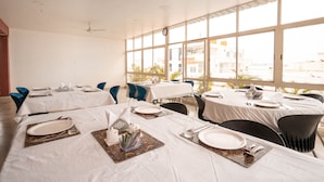 Un comedor con varias mesas dispuestas para comer, ventanas amplias con vista a edificios y ventiladores de techo.