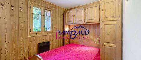 A bedroom with wooden walls, a bed with a pink bedspread, a window, and wooden cabinets.