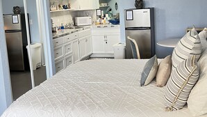A bedroom with a large bed, a small desk, and a kitchenette in the background. - ~ Shore ~ Thing ~ Studio Condo near The Beach ~ (Daytona Beach)