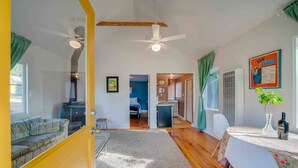 Une pièce lumineuse avec une porte jaune, un ventilateur de plafond, une table à manger ornée d’un vase et d’une bouteille, et une vue sur une autre pièce où se trouvent un lit et un bureau.