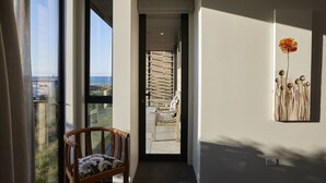 Un interior moderno con vista al mar a través de un ventanal grande, una silla de madera y un arreglo floral colgando en la pared.
