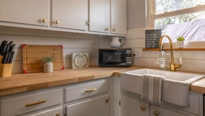 A kitchen with white cabinets, a wooden countertop, a microwave, a sink with a gold faucet, and a window with a view of trees. - Charming open 4bd and screen-in porch! Sleeps 10! (Saint Charles)