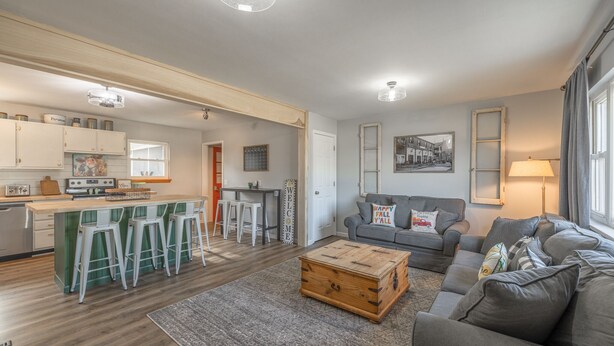 A spacious living area with a grey sofa, a wooden coffee table, and a kitchen area with a bar stool seating. - Charming open 4bd and screen-in porch! Sleeps 10! (Saint Charles)