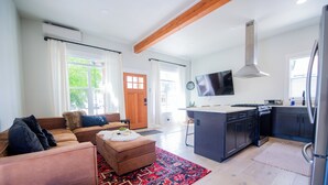 Una sala de estar moderna con un sofá, un reposapiés y una alfombra estampada. Se ve una cocina al fondo con estufa, fregadero y refrigerador.
