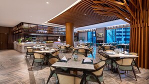 Ein geräumiges Restaurant mit hohen Decken, Holzbalken und einer zentralen Bar. Tische sind mit weißen Tischdecken und Korbstühle gedeckt.