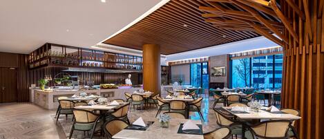 A spacious restaurant with high ceilings, wooden beams, and a central bar area. Tables are set with white tablecloths and wicker chairs.