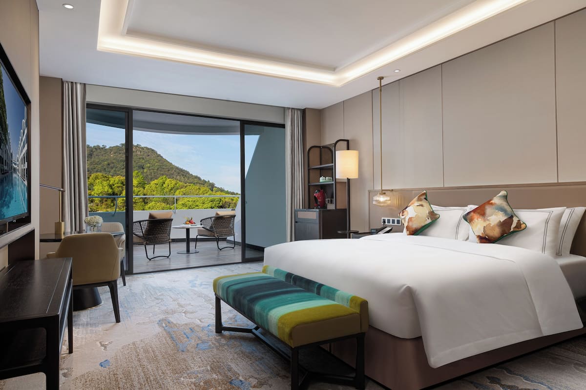 A hotel room with a large bed, a desk, a chair, and a bench. There is a balcony with a view of greenery.