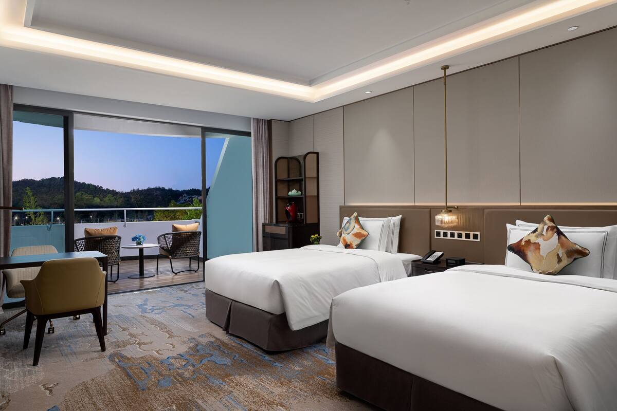 A hotel room with two beds, a desk, a chair, and a view of the outdoors.