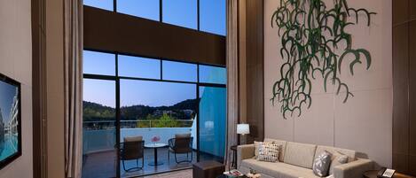 A modern hotel room with a large sliding glass door leading to a balcony, a sofa set, a small table, and a decorative wall piece.