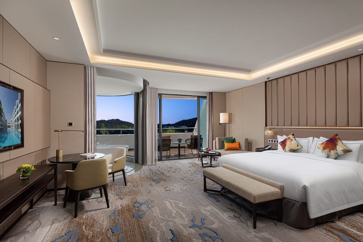 A modern hotel room with a large bed, a desk, a chair, and a balcony with a view.