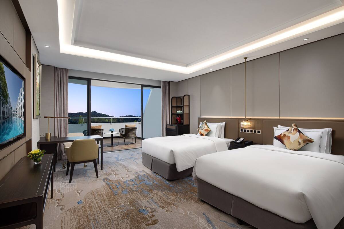 A hotel room with two beds, a desk, a chair, a large window, and a balcony.
