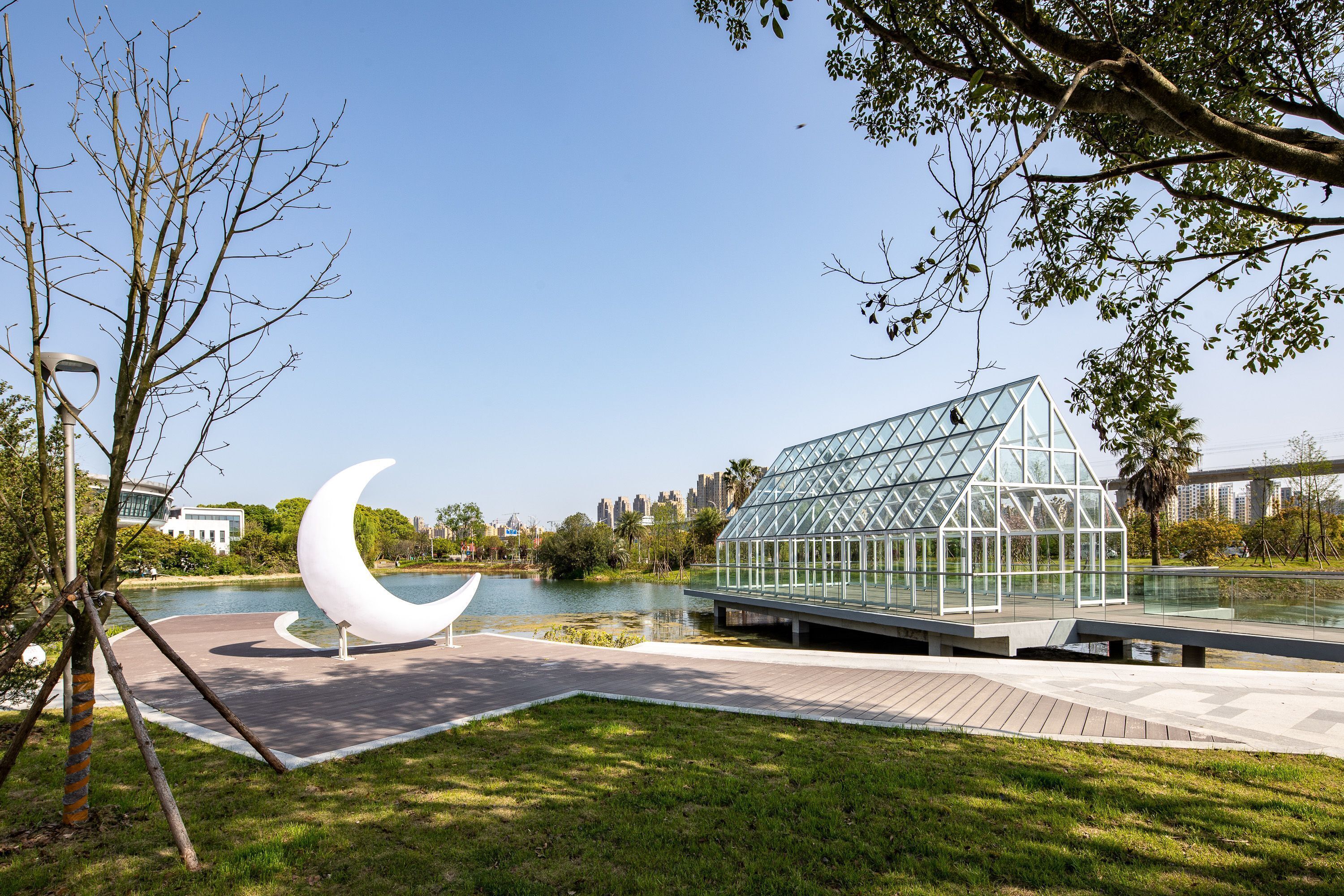 A park with a glass greenhouse structure, a crescent moon sculpture, and a lake.