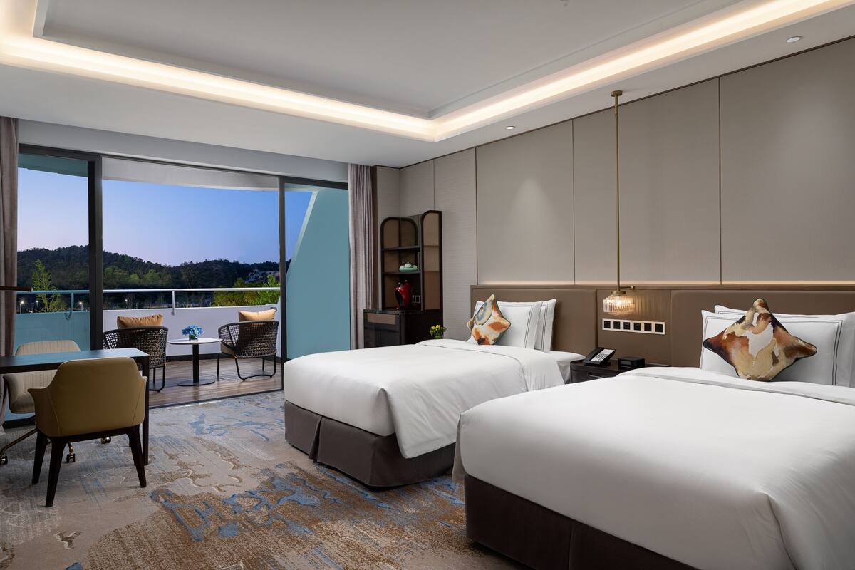 A hotel room with two beds, a desk, and a balcony with a view of the hills.