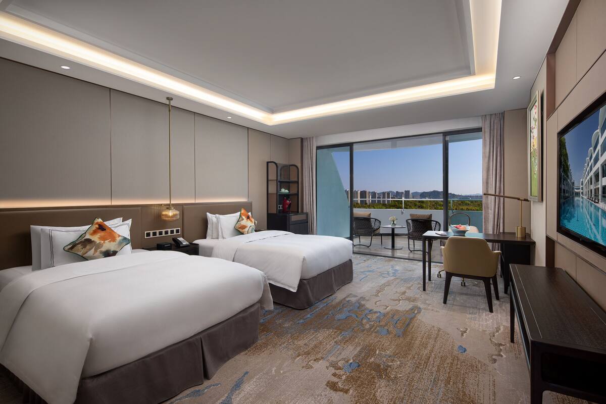 A hotel room with two beds, a desk, a chair, a TV, and a balcony with a city view.