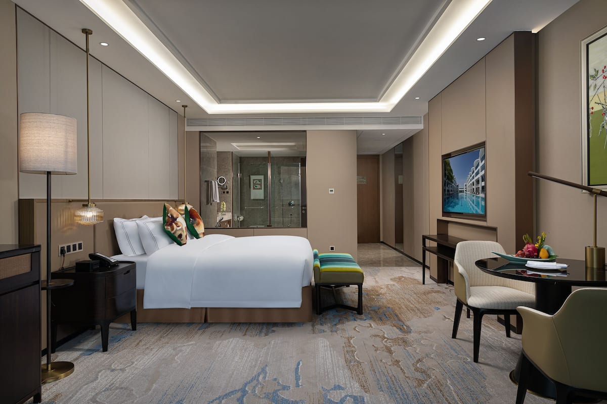 A modern hotel room with a large bed, bedside tables, a desk with a chair, a small stool, and a television.