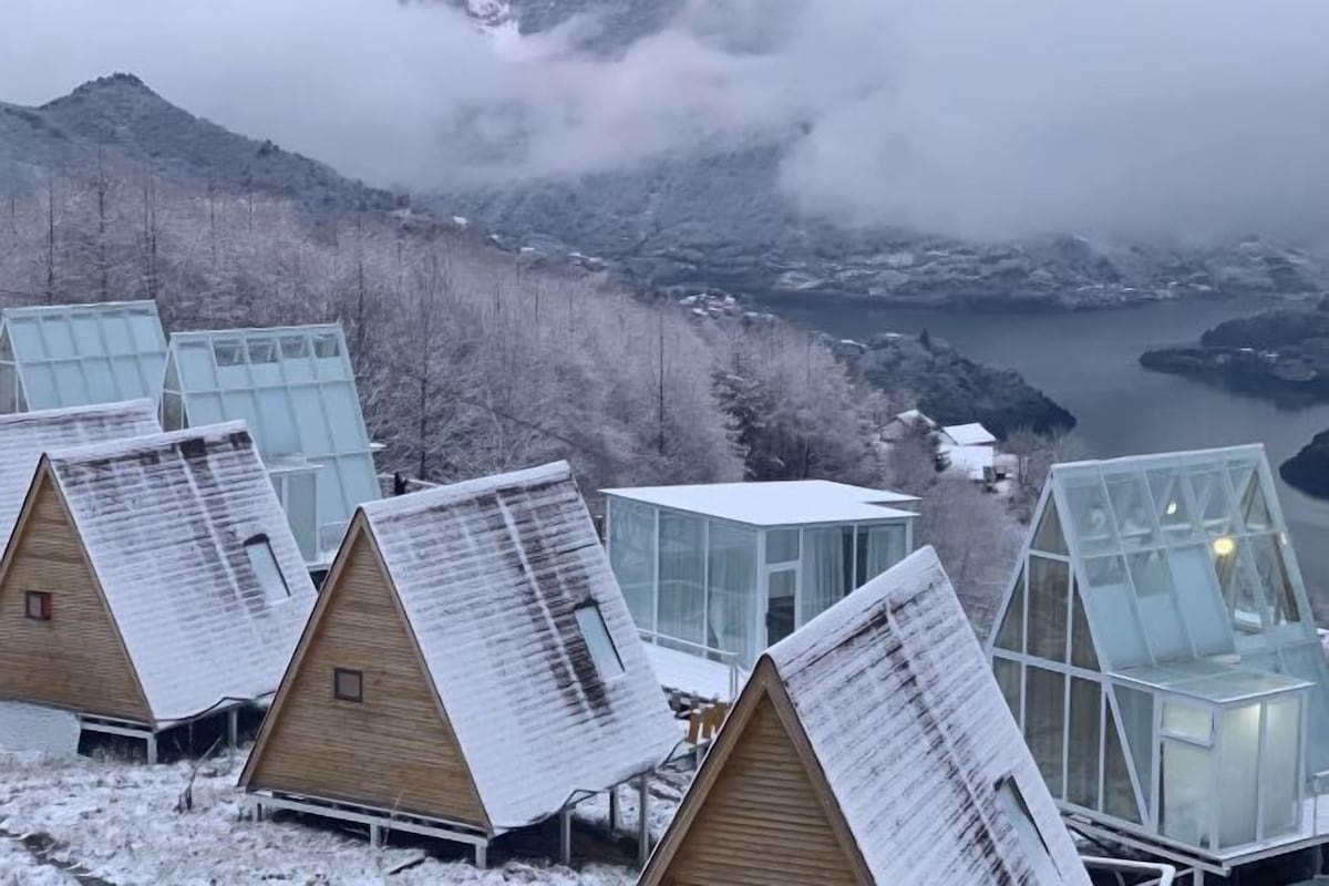 Eine Reihe von Holzhütten mit Glasdächern vor einer Berglandschaft und einem Gewässer.