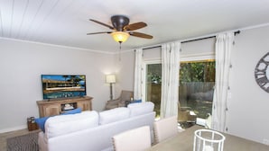 Ein Wohnzimmer mit einer Couch, einem Fernseher, einem Deckenventilator, einer Lampe und einem großen Fenster mit Vorhängen.
