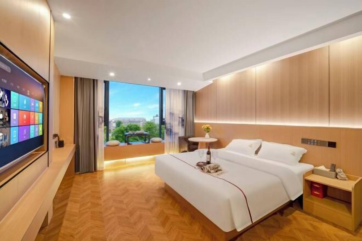 Een moderne hotelkamer met een groot bed, een televisie aan de muur, een tafeltje met een vaas, en uitzicht op de stad.